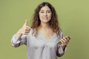 smiling-young-pretty-female-office-worker-holding-phone-her-thumb-up-isolated-olive-green-wall-min smiling-young-pretty-female-office-worker-holding-phone-her-thumb-up-isolated-olive-green-wall-min